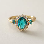 Oval teal blue center stone surrounded by sparkling cubic zirconia cz crystal halo three stone band stainless steel jewelry ring in waterproof 18k gold plated finish.