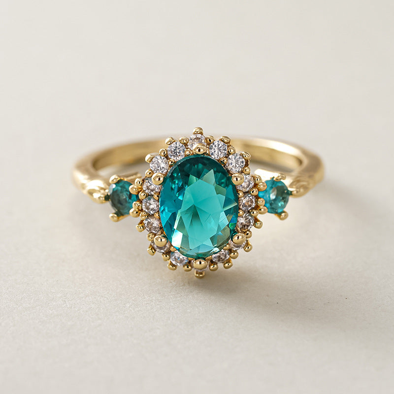 Oval teal blue center stone surrounded by sparkling cubic zirconia cz crystal halo three stone band stainless steel jewelry ring in waterproof 18k gold plated finish.