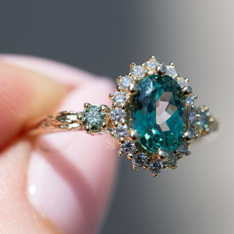 Oval teal blue center stone surrounded by sparkling cubic zirconia cz crystal halo three stone band stainless steel jewelry ring in waterproof 18k gold plated finish.