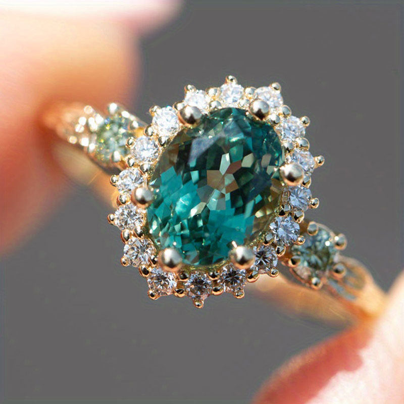 Oval teal blue center stone surrounded by sparkling cubic zirconia cz crystal halo three stone band stainless steel jewelry ring in waterproof 18k gold plated finish.