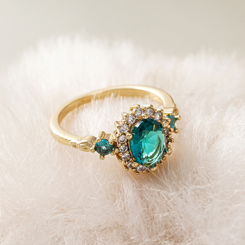 Oval teal blue center stone surrounded by sparkling cubic zirconia cz crystal halo three stone band stainless steel jewelry ring in waterproof 18k gold plated finish.